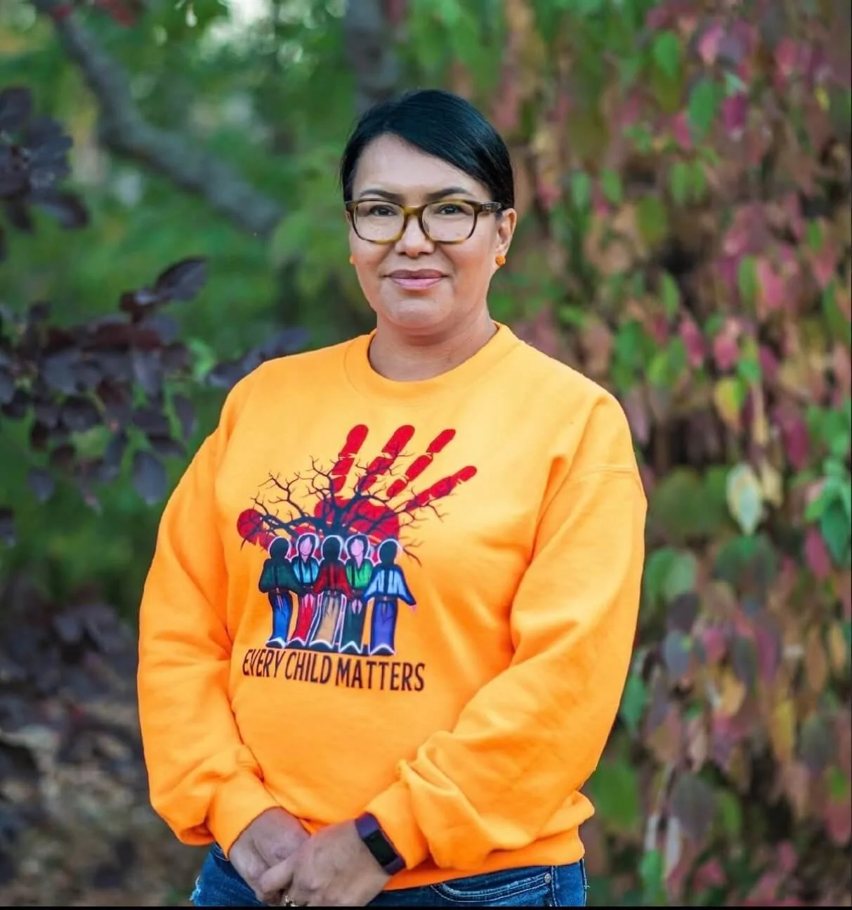 Brenda Dreaver wearing glasses and an orange sweatshirt with a graphic design of four diverse women and a tree with roots, along with the text "Every Child Matters." She stands outdoors in front of colourful autumn foliage.