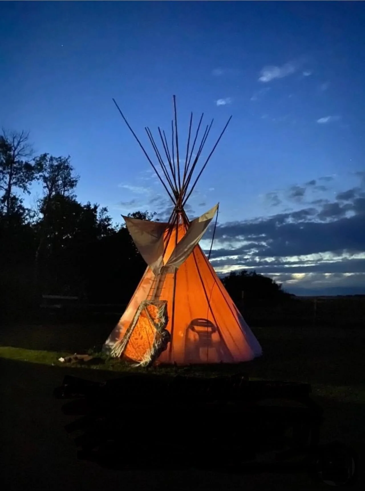 A glowing teepee tent illuminated from inside, set against a twilight sky with silhouetted trees and clouds.