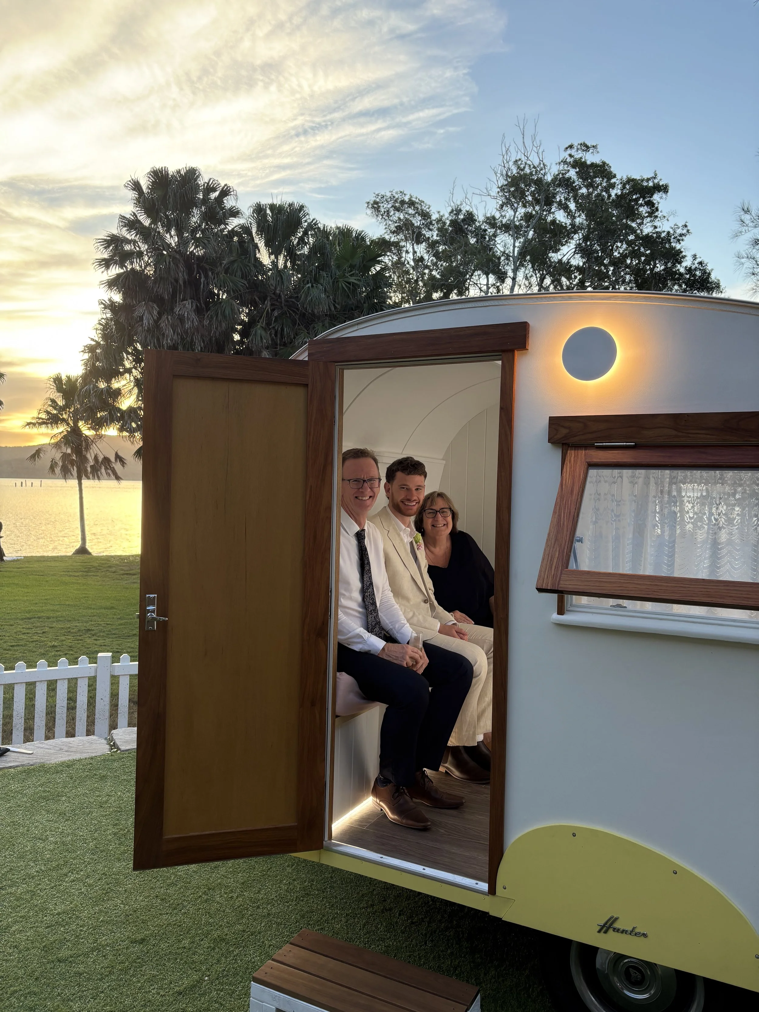 Three people sitting inside a tiny house on wheels by a lake during sunset, smiling at the camera.