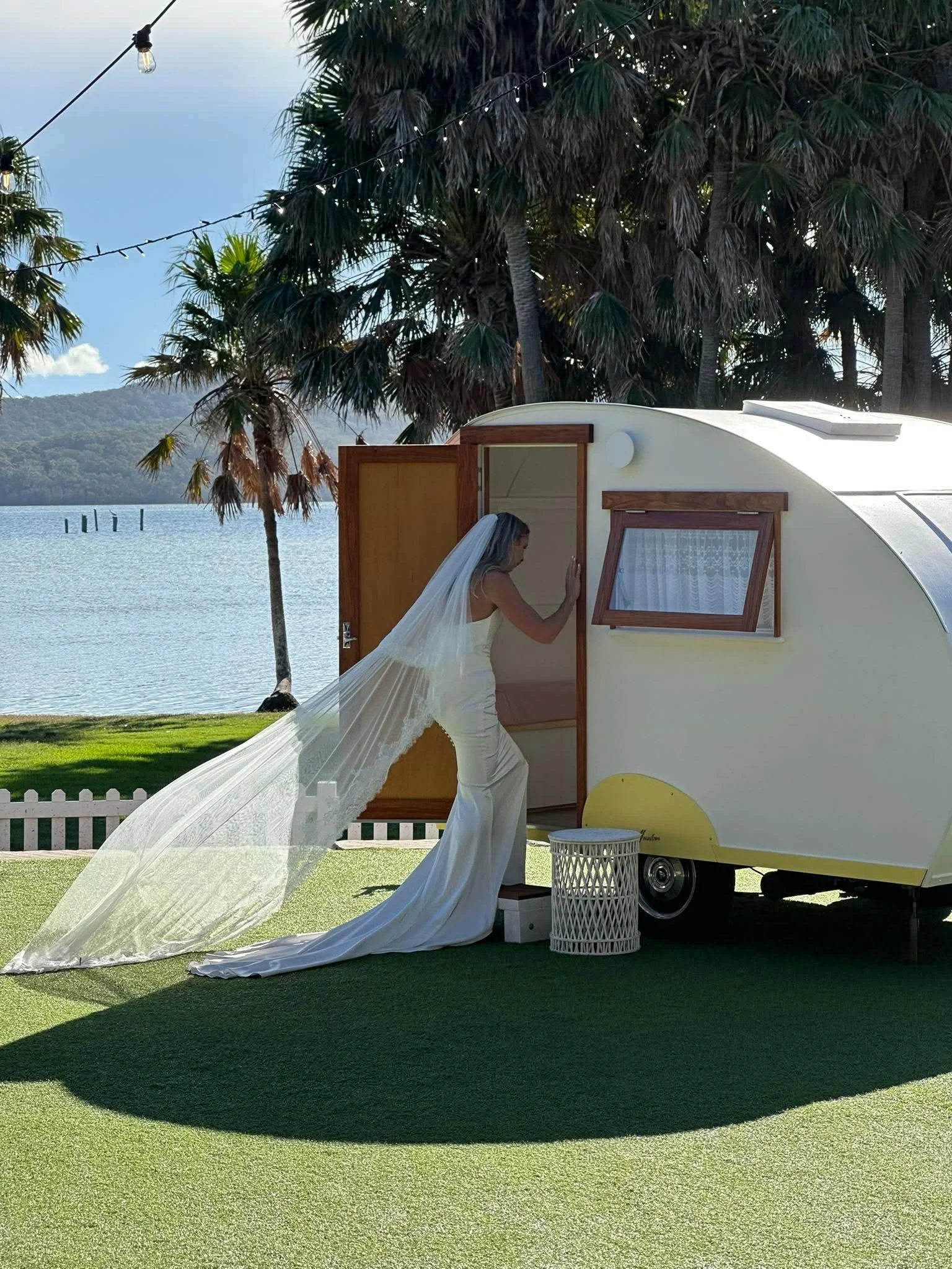 Brunette woman in wedding dress with long veil standing outside a tiny travel trailer near a lake with palm trees.