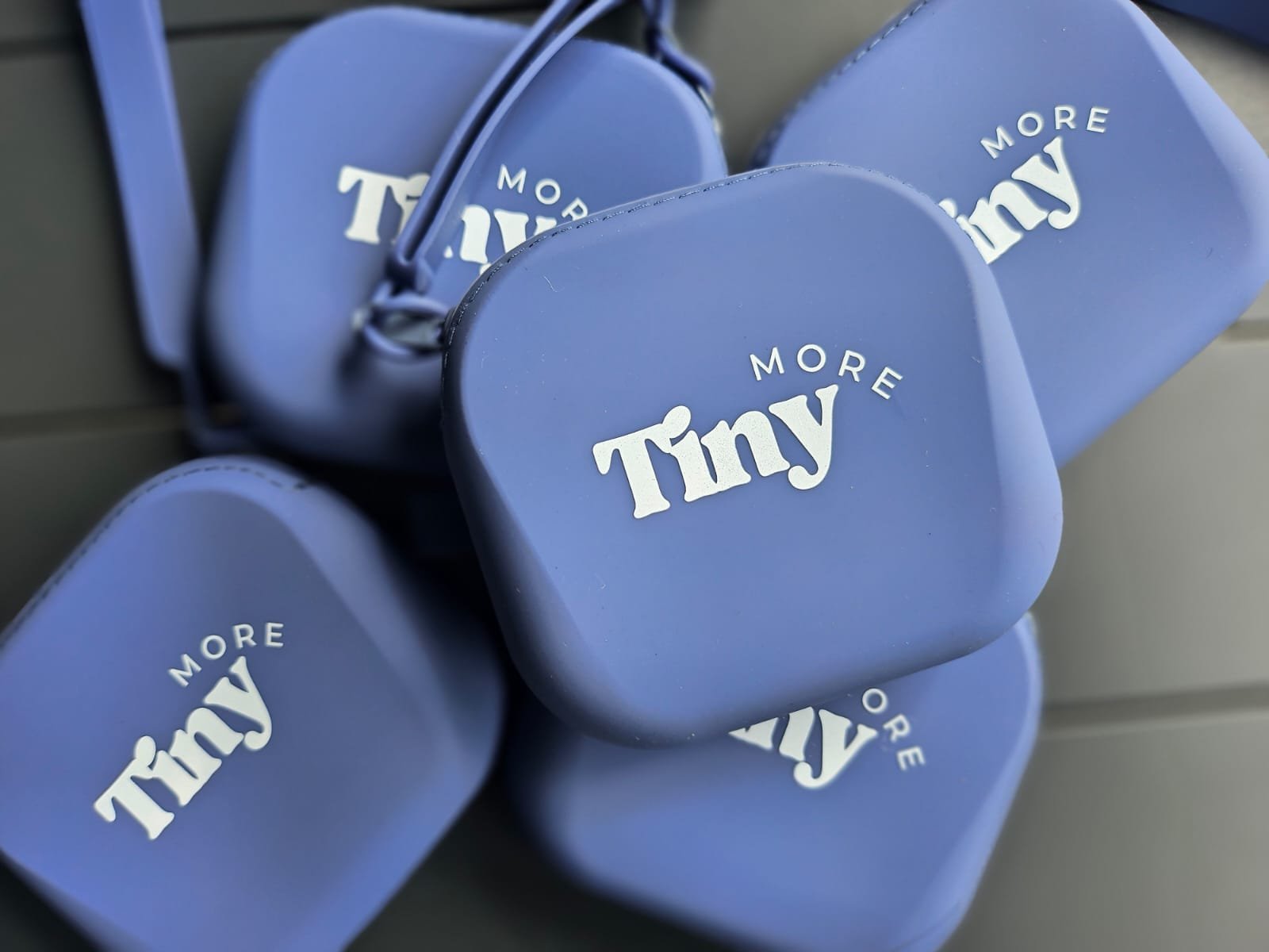Multiple blue cases with white printed text that reads 'Tiny More'. 	Reusable and splash-proof silicone containers showing carry handle and quality of pouch to hold condiments.