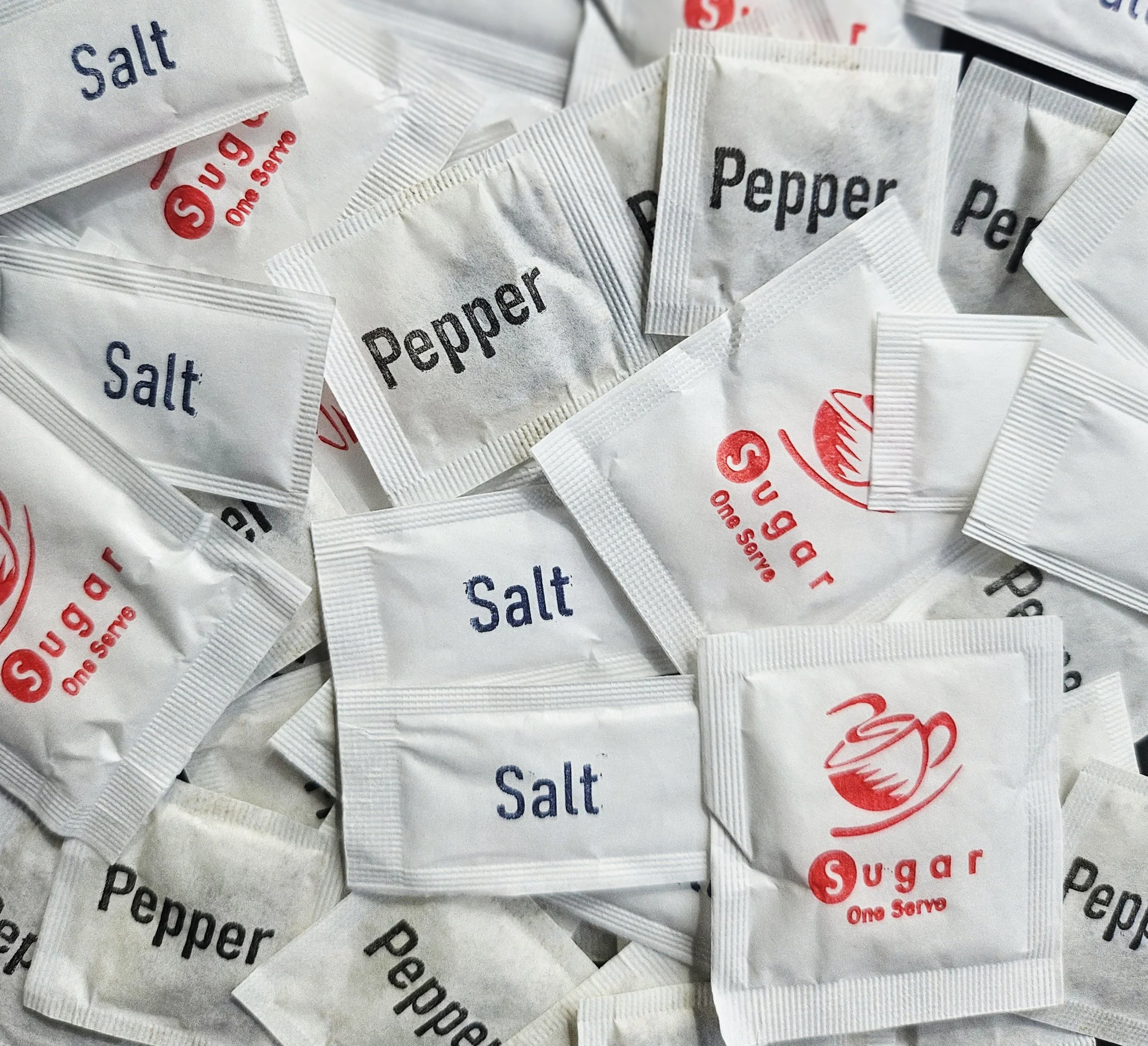 A pile of sugar and salt packets, with some labeled 'Salt' in blue and others labeled 'Sugar' in red, some with the phrase 'One Serve' and a cup icon. These items that are included in 'Tiny More' packs.