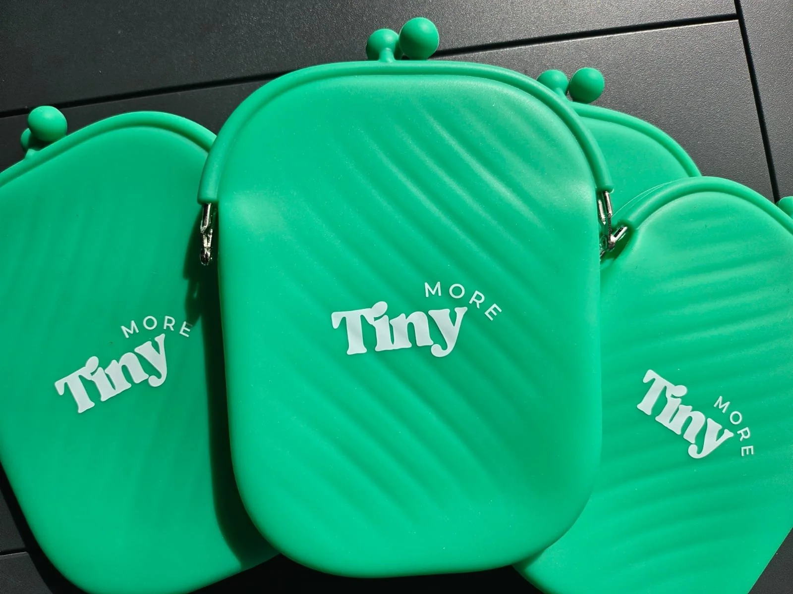 Three green coin purses with the words "Tiny More" printed on them in white text. Showing quality silicone material that is splash-proof and can be re-purposed.