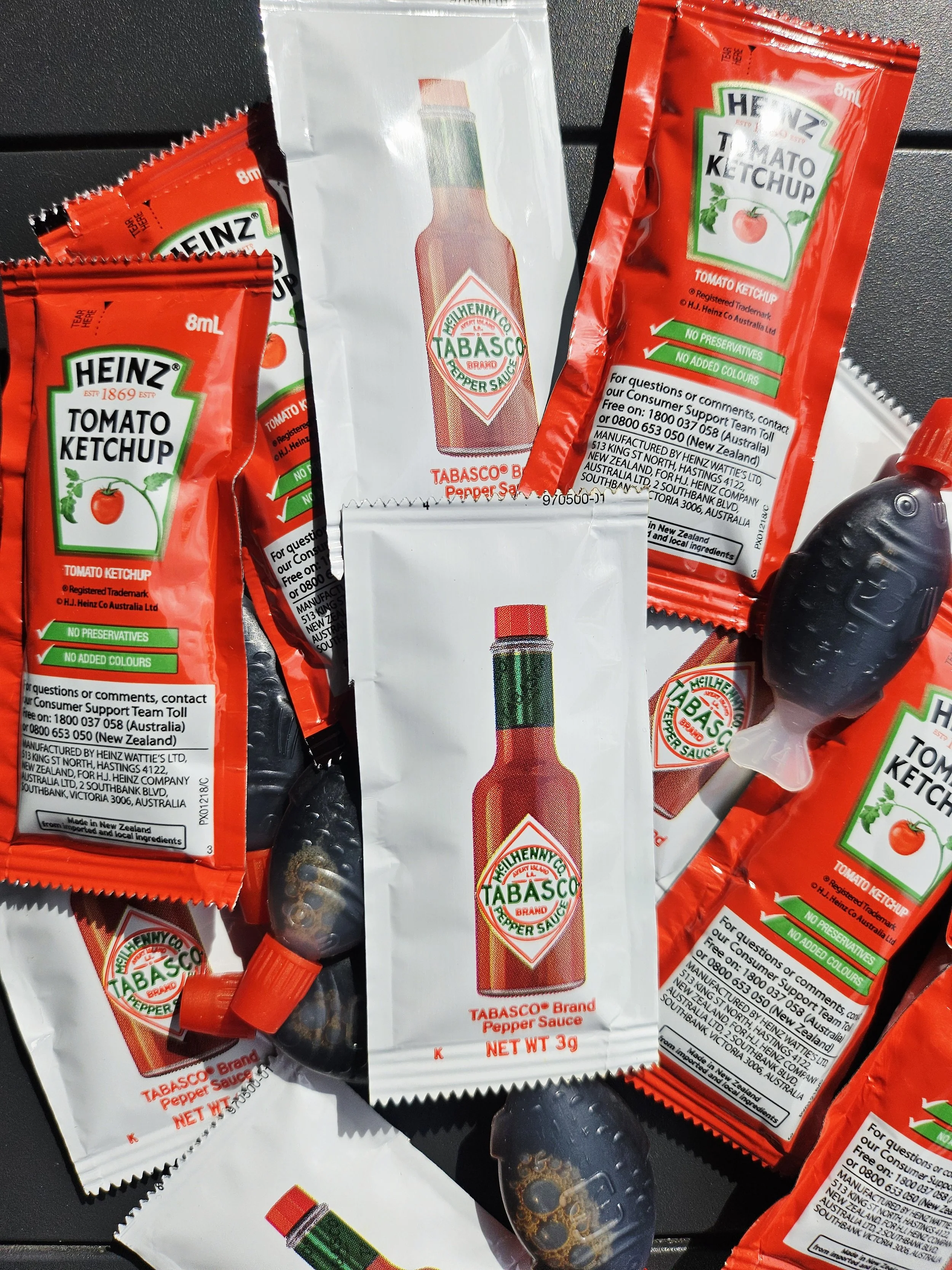 Multiple small packets of Heinz tomato ketchup with red packaging, a packet of Tabasco pepper sauce with a red cap, and black plastic Soy sauce squeeze bottles with black caps on a dark surface. These items that are included in 'Tiny More' packs.