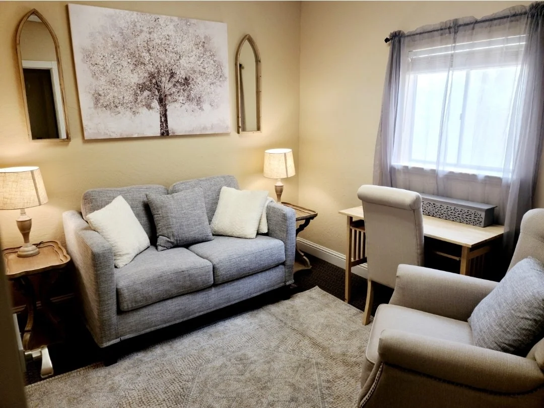 Living room with a grey sofa, white armchair, beige armchair, two table lamps, wall art of a tree, window with sheer curtains, and a small desk.
