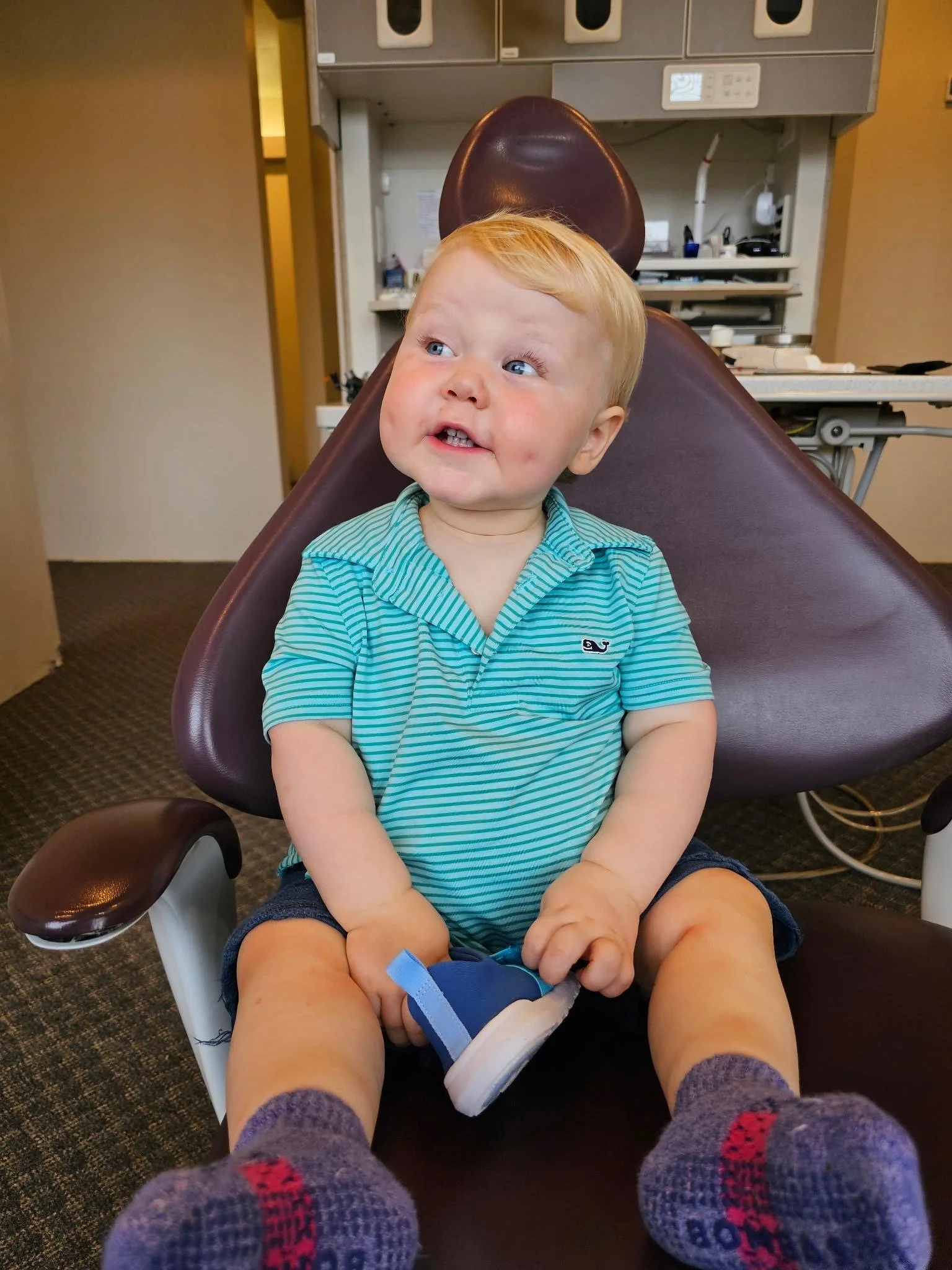 A young child with blond hair, blue eyes, and a striped turquoise shirt sitting in a dental chair, looking to the side with a smile, holding a blue shoe, with a background of dental equipment.