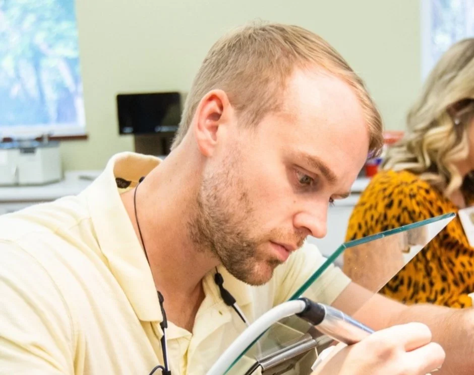 A man in a yellow shirt is reading or writing on a notepad, with a glass partition in front of him, in a classroom or office setting.