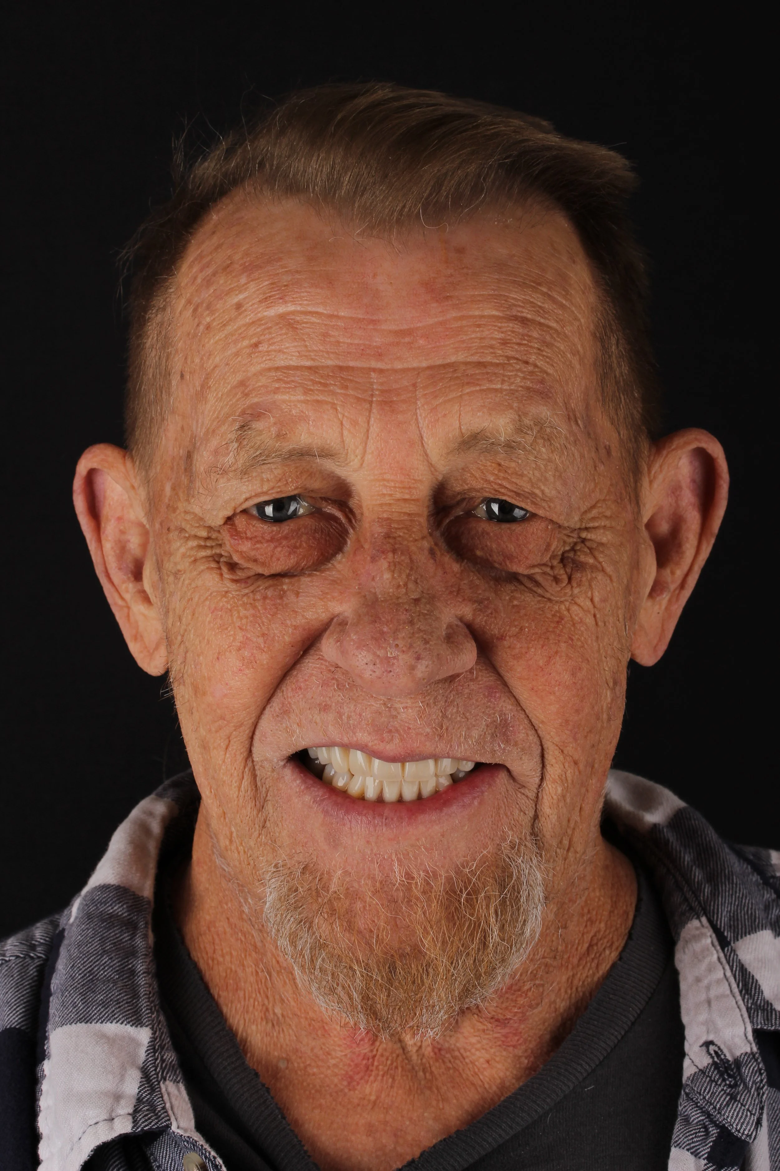 Close-up of an elderly man with a wrinkled face, gray hair, and a goatee, smiling with some teeth visible and one eye partially closed, against a black background.