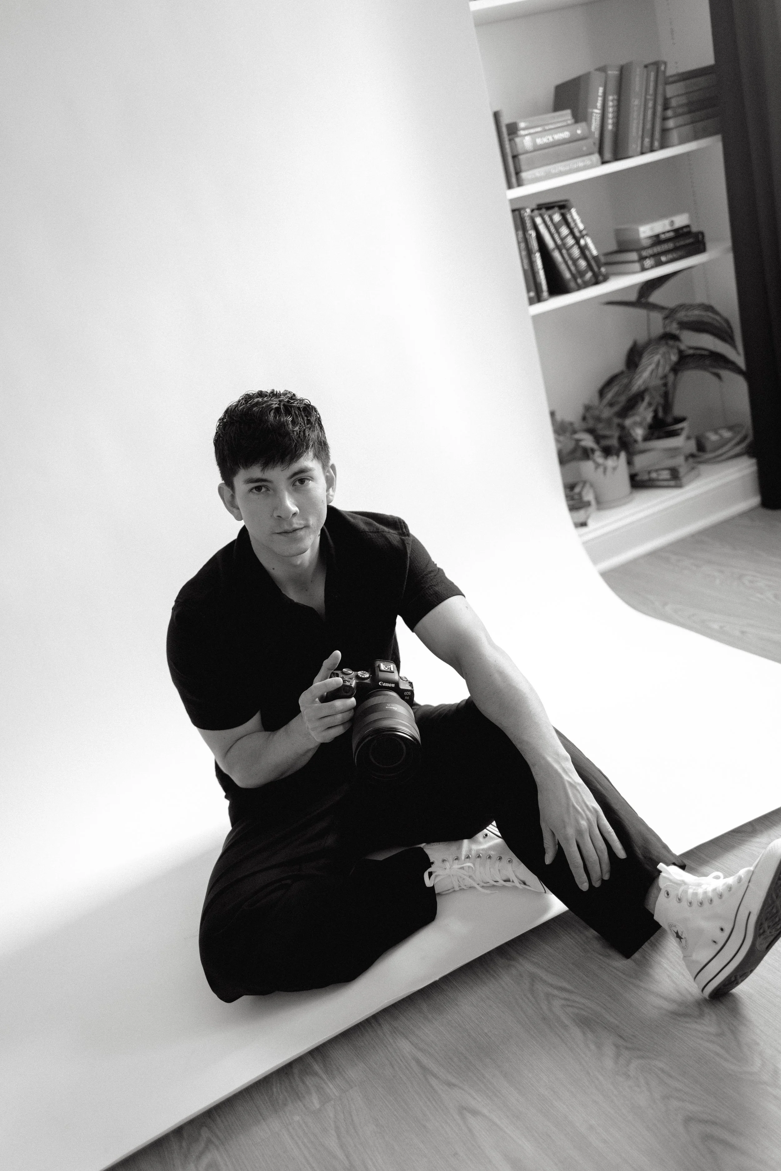 A young man with short dark hair, wearing a black shirt, black pants, and white sneakers, sits on the floor in front of a white backdrop, holding a camera. There are books and a potted plant on a bookshelf in the background.