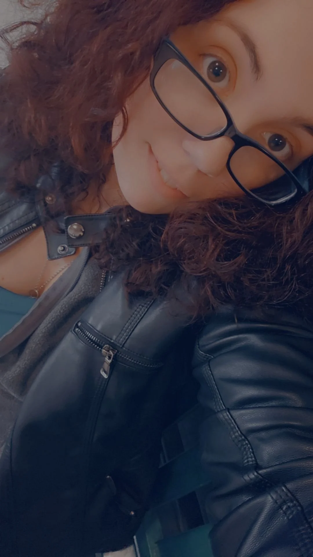 A woman with curly hair and glasses smiling, wearing a black leather jacket.