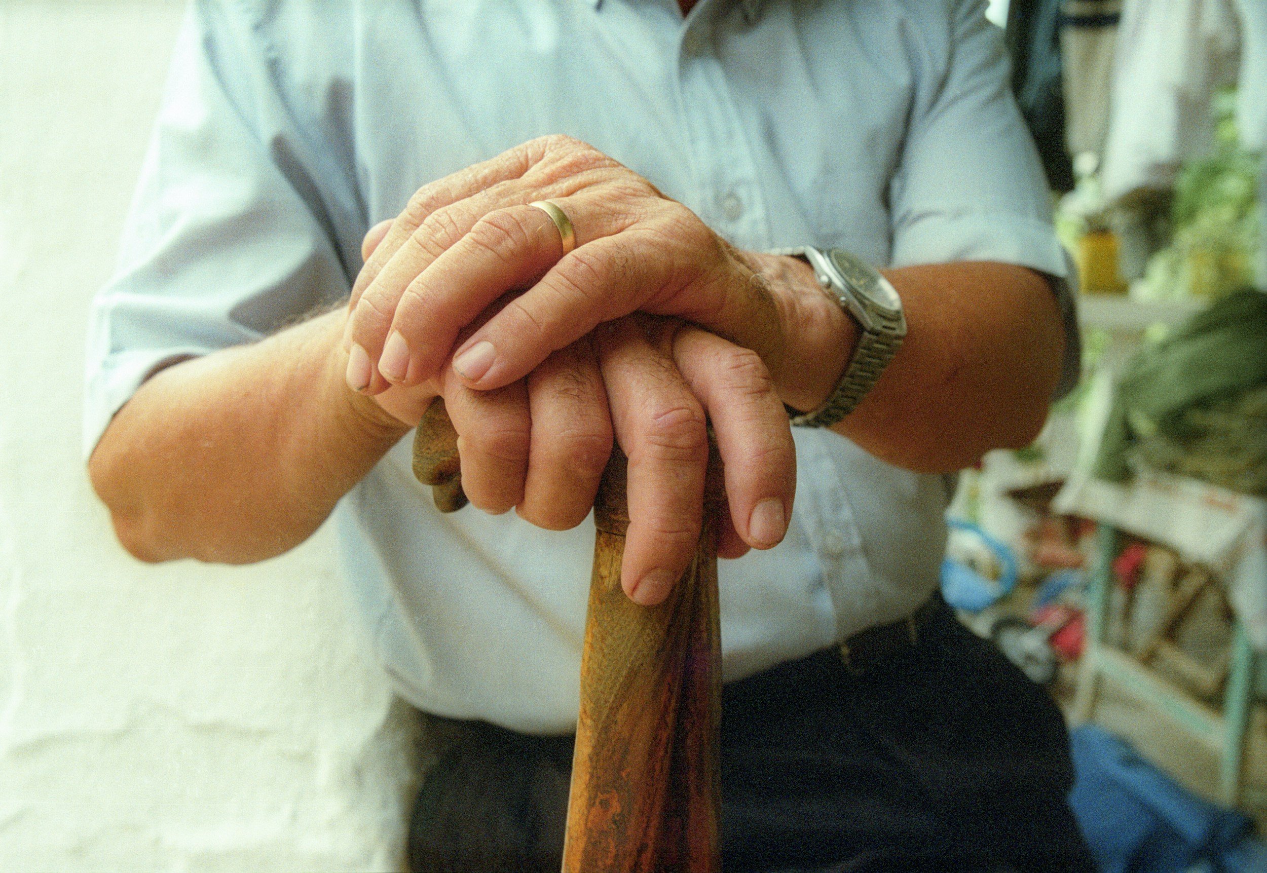A person clasping hands with an elderly individual, using a wooden walking cane, with shelves of colorful items in the background.