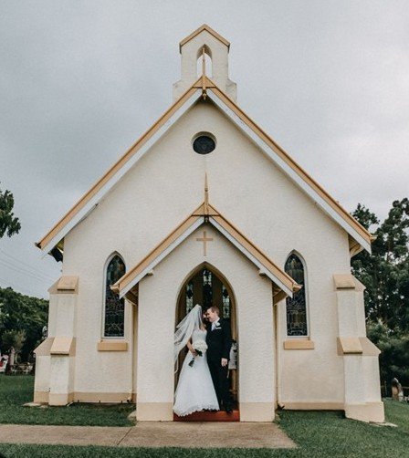 A bride and groom standing in front of a small beige church with a steeple and arched windows on a cloudy day.