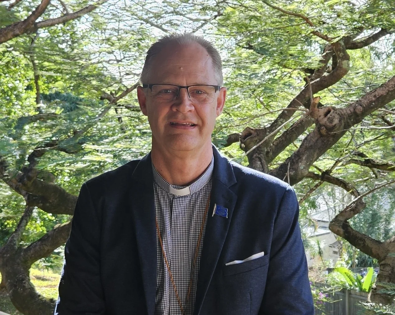A man dressed as a bishop, wearing a clerical collar and glasses, standing outdoors in front of a leafy tree background.