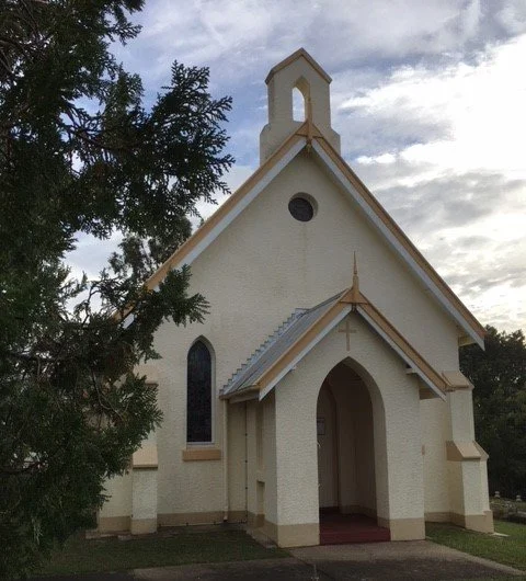A small white church with a pointed roof, arched entrance, and a small bell tower on top, surrounded by trees under a cloudy sky.