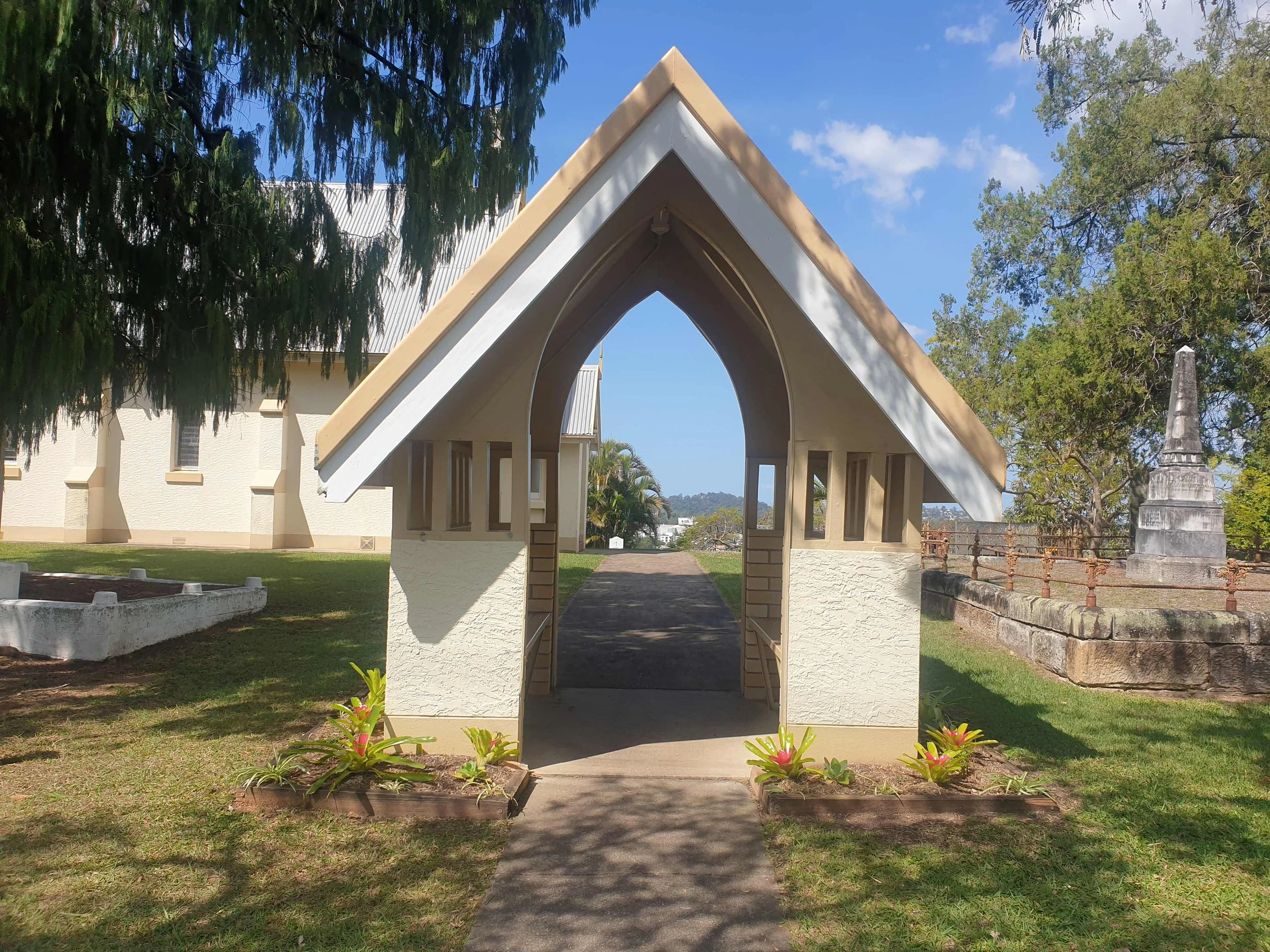 Small white chapel with a gabled roof, surrounded by grass, trees, and a monument on a stone base.