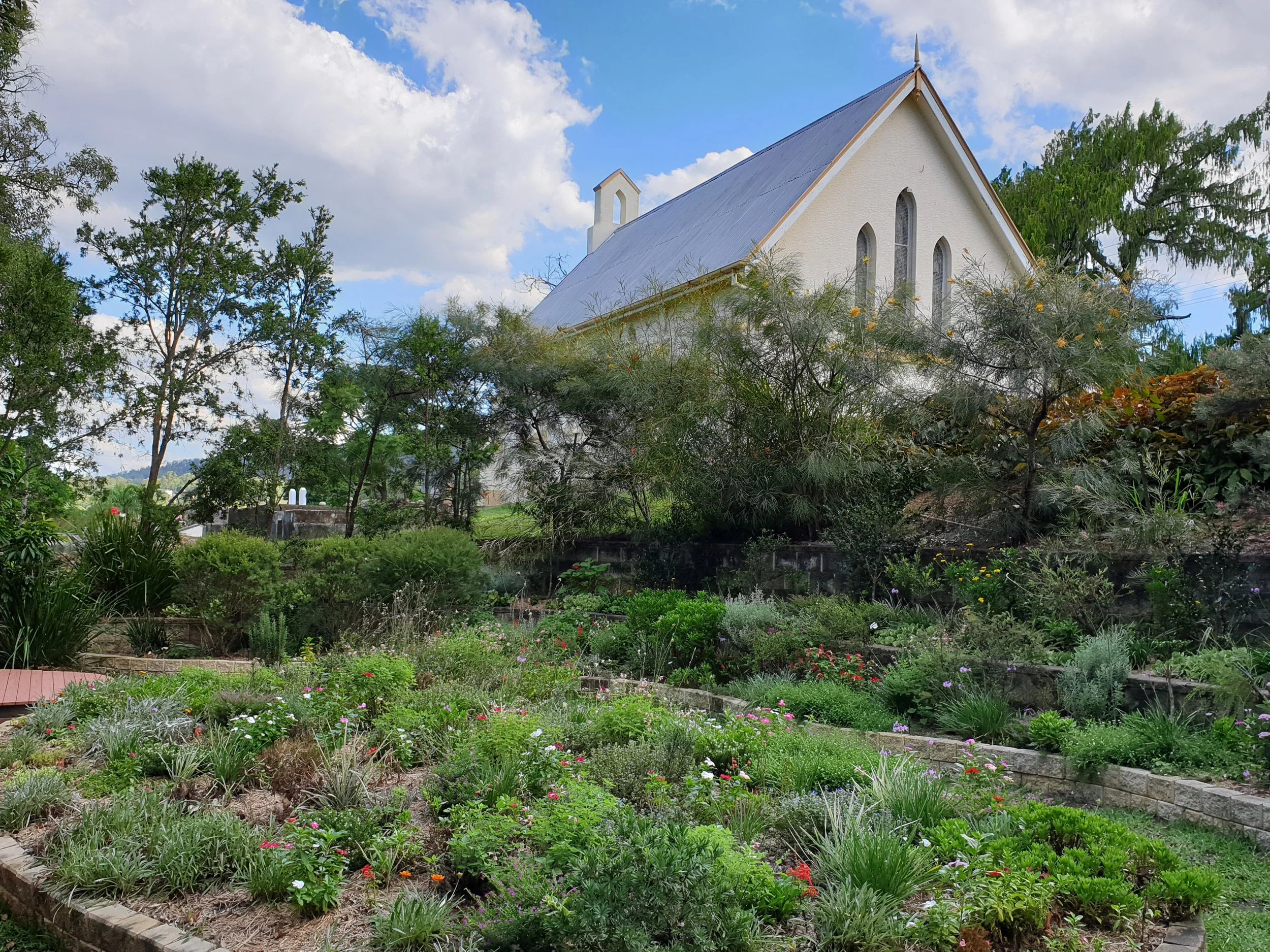 A white church with a steep roof among green trees and a garden with colorful flowers and bushes.
