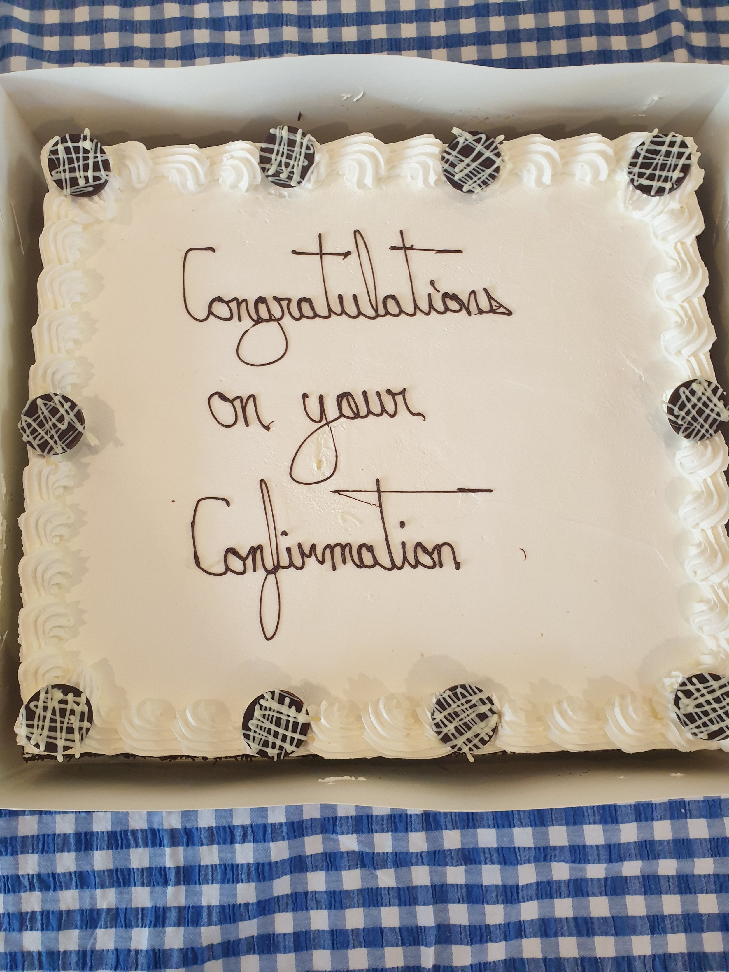 A rectangular white cake with whipped frosting, decorated with chocolate truffles drizzled with white chocolate on top and around the edges, on a table with a blue and white checkered tablecloth. The cake has the message 'Congratulations on your Congratulations' written in cursive icing.