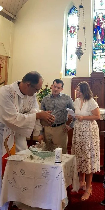 A man dressed as a priest or minister performs a religious ceremony with two people, a man and a woman, in a church with stained glass windows.