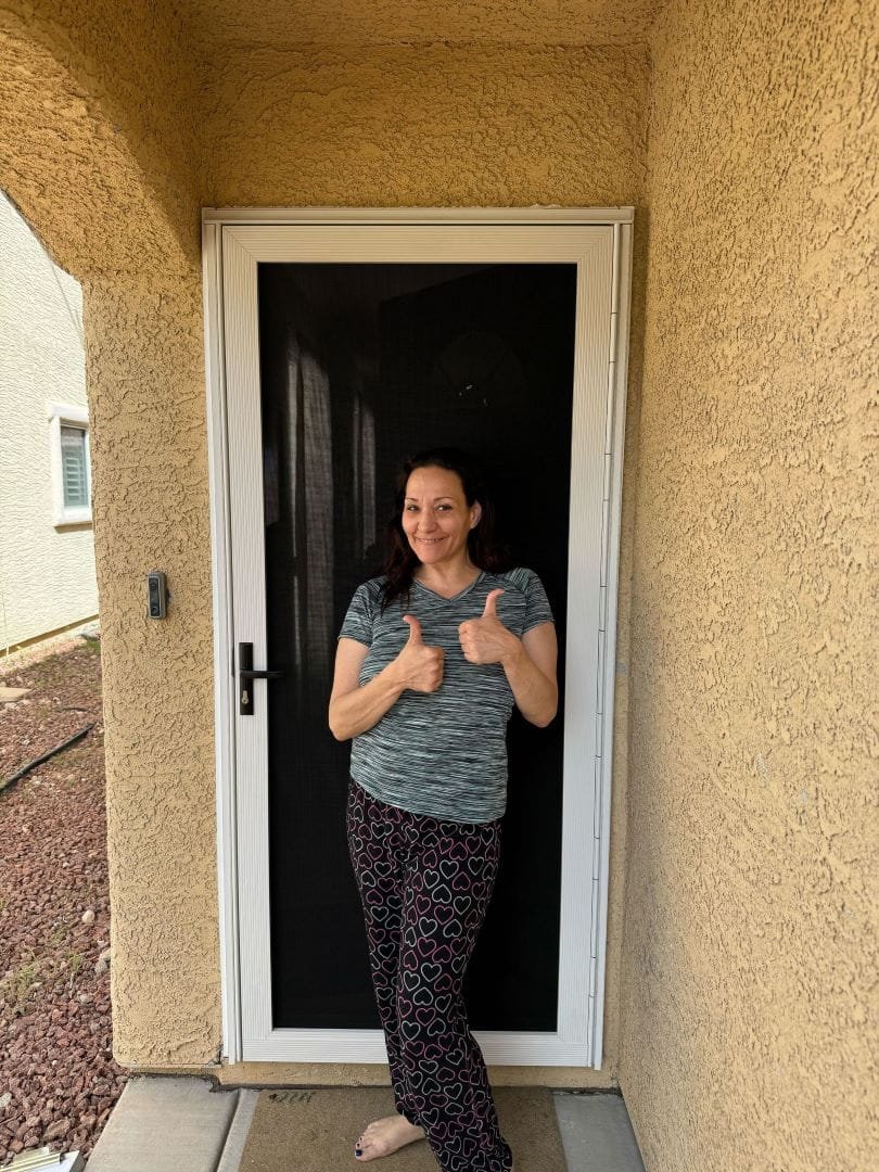 A woman smiles and gives two thumbs-up while standing in her doorway behind a new black security screen door.