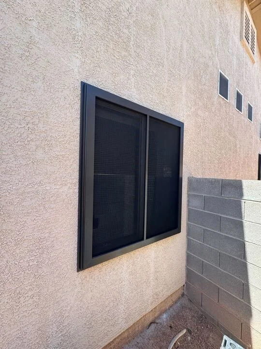 An exterior side view of a house showing a window with a thick black frame and a dark sun screen, set into a light-colored stucco wall. A gray block wall is visible in the foreground.