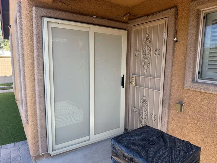 An exterior shot of a tan-framed sliding screen door and an ornate metal security door installed side-by-side on the back of a stucco house.