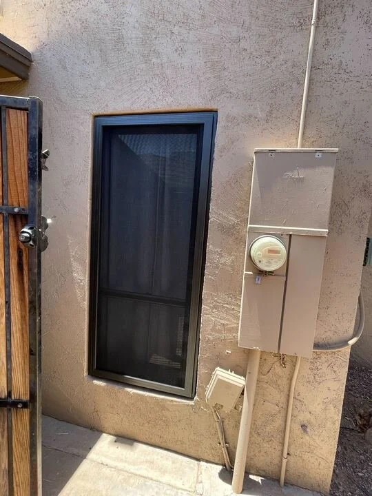A tall, black-framed security screen door installed in a narrow walkway on the side of a stucco house, next to a utility meter box.