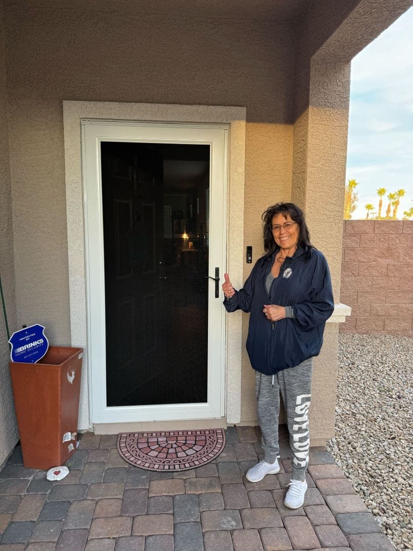A smiling woman stands on her front porch, presenting her new white-framed security screen door with black mesh.