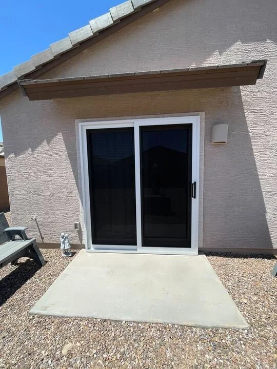 An exterior view of a white-framed sliding glass door with dark sun screens installed on a tan stucco house. The door opens onto a concrete patio surrounded by gravel.