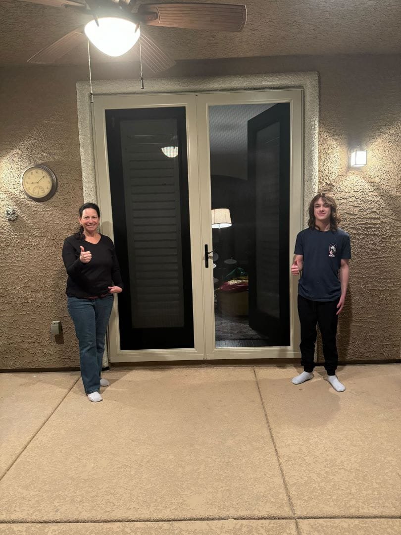 A woman and a young man stand on a covered patio, giving a thumbs-up next to a newly installed set of beige-framed screen doors.