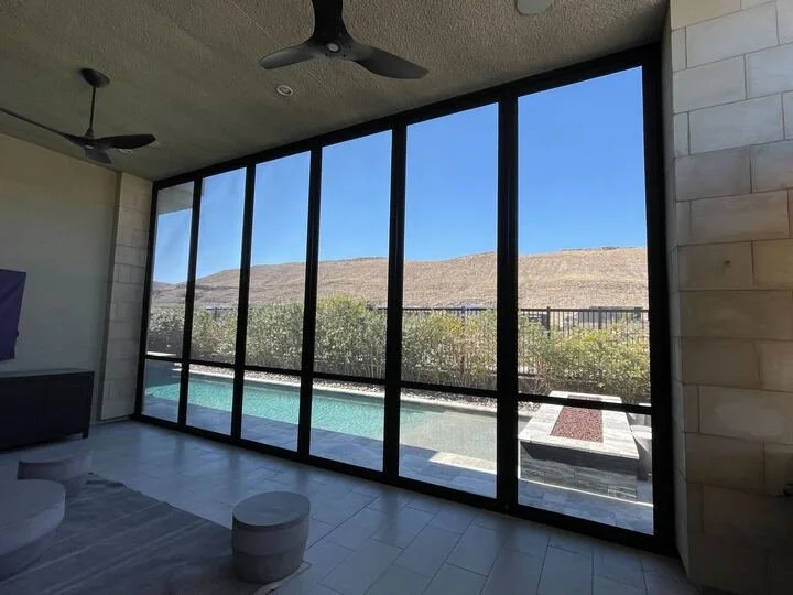 An interior shot of a room with a large wall of black-framed windows looking out onto a backyard with a swimming pool and a desert hillside under a clear blue sky. Two ceiling fans are visible on the ceiling.