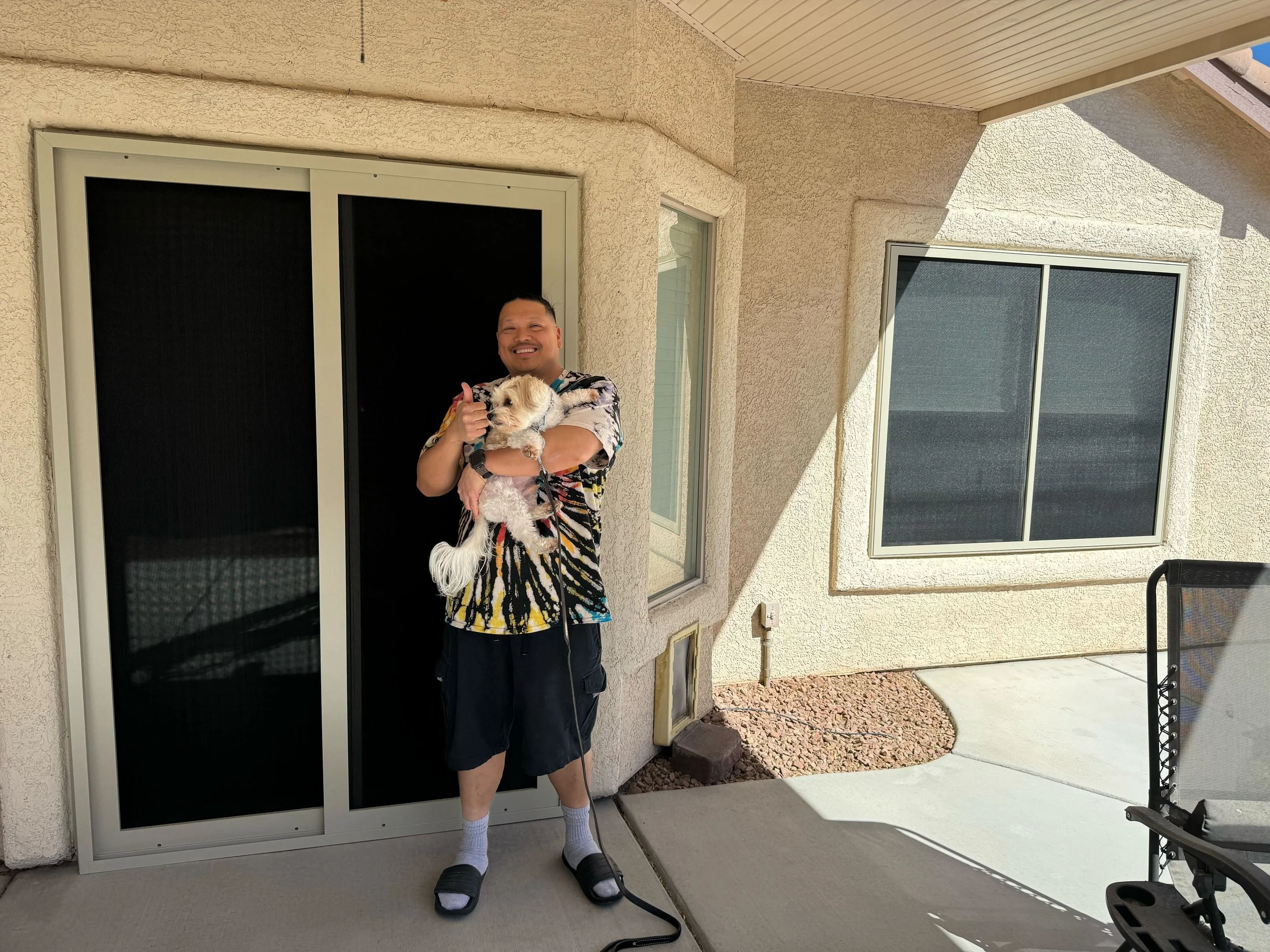 A man holding a small, fluffy dog stands on a concrete patio in front of a sliding glass door fitted with new, dark sun screens.