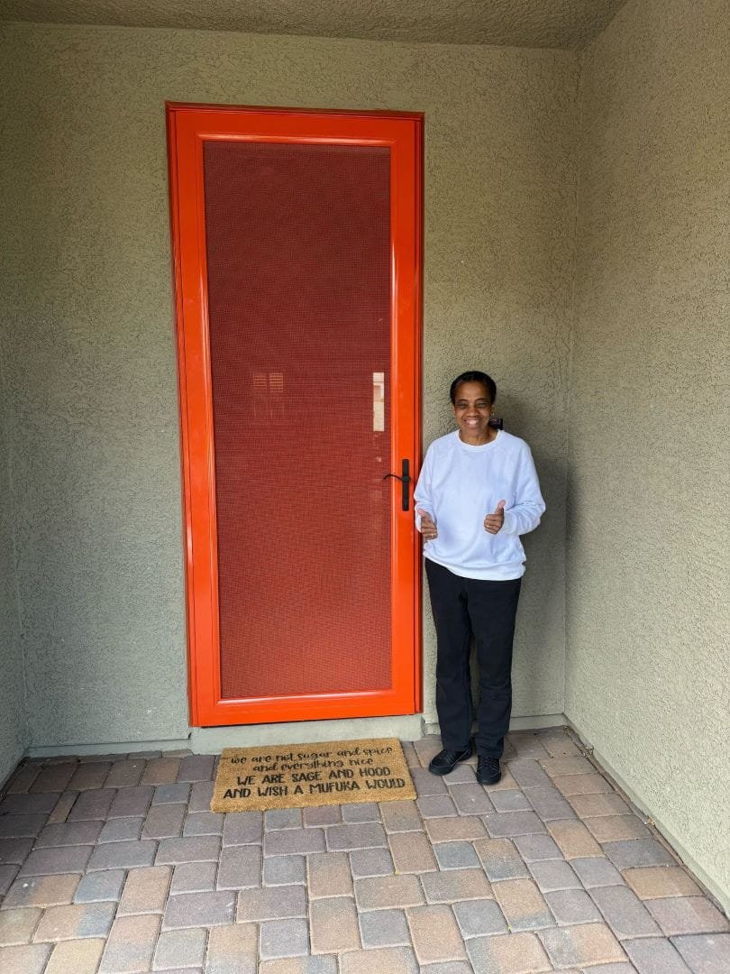 A woman smiles with two thumbs-up while standing in her entryway next to a new, bright orange-framed security screen door.
