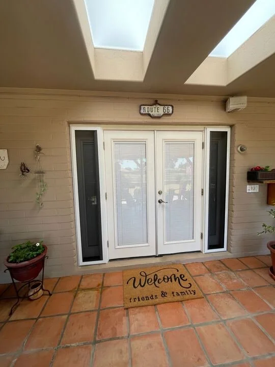 The front entryway of a house, showing white double doors, a "Welcome friends & family" doormat on a terra-cotta tile floor, and two skylights overhead.