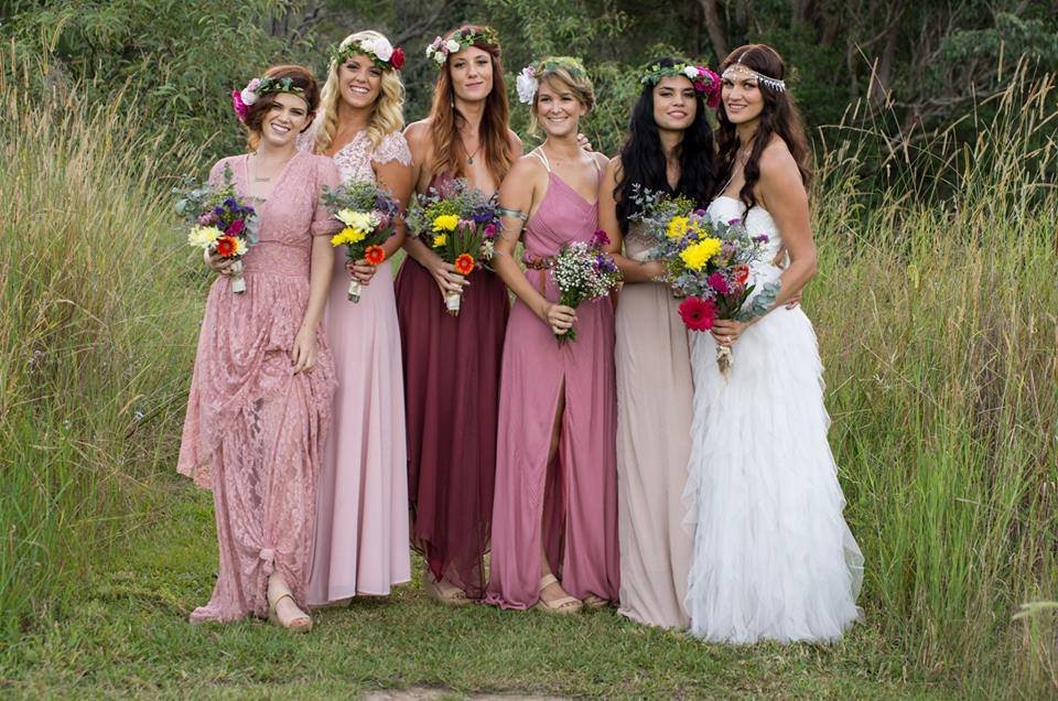 Six women in colorful dresses holding bouquets of flowers standing in a grassy outdoor area.