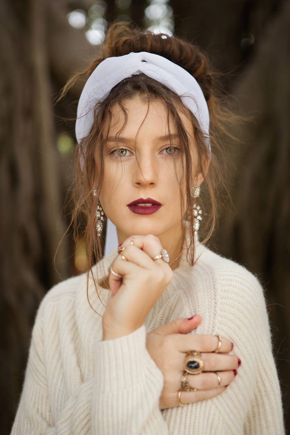 A woman with red hair, styled in an updo with a white headband, wearing jewelry including rings, earrings, and a necklace, standing outdoors in front of tree trunks with bokeh background, looking directly at the camera.