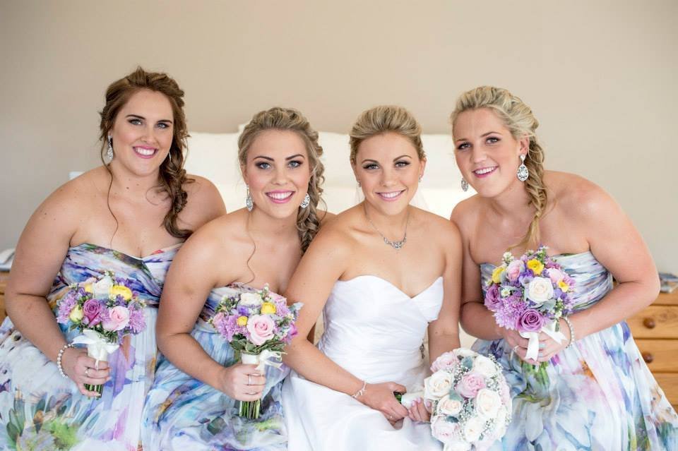 Four women, including a bride and bridesmaids, smiling and holding bouquets, dressed in wedding attire, in a room with beige walls.