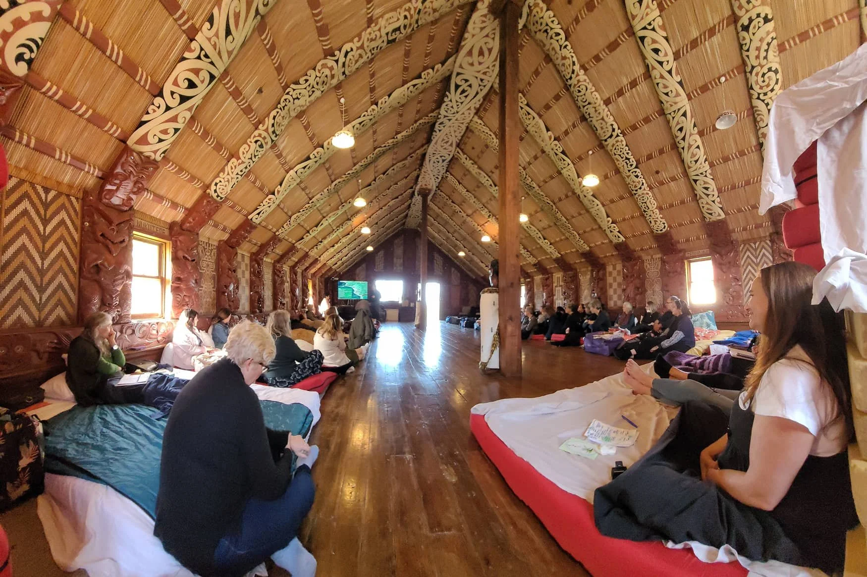 People sitting on mattresses and cushions in a large, decorated hall with wooden floors and walls, attending a gathering or meditation session.