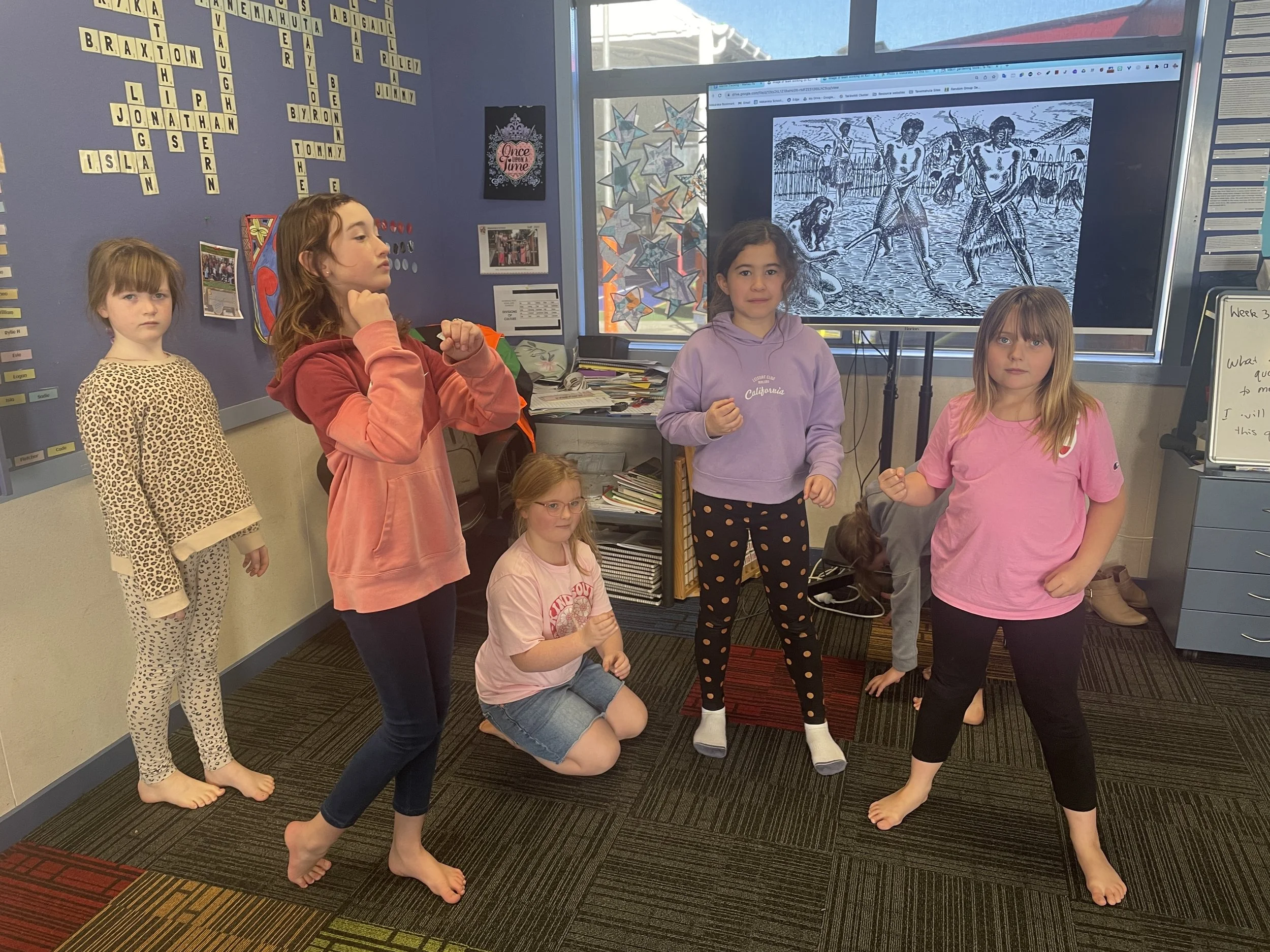 A group of six students gathered in a classroom, standing and kneeling in front of a large screen displaying a Maori gardening scene.