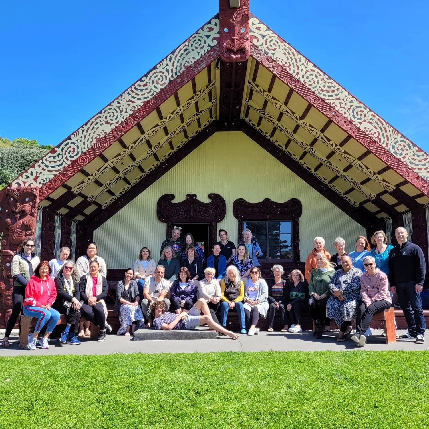 A group of people gathered in front of a traditional Maori meeting house with intricate carvings and a rafter with a carved face at the peak, under a clear blue sky.