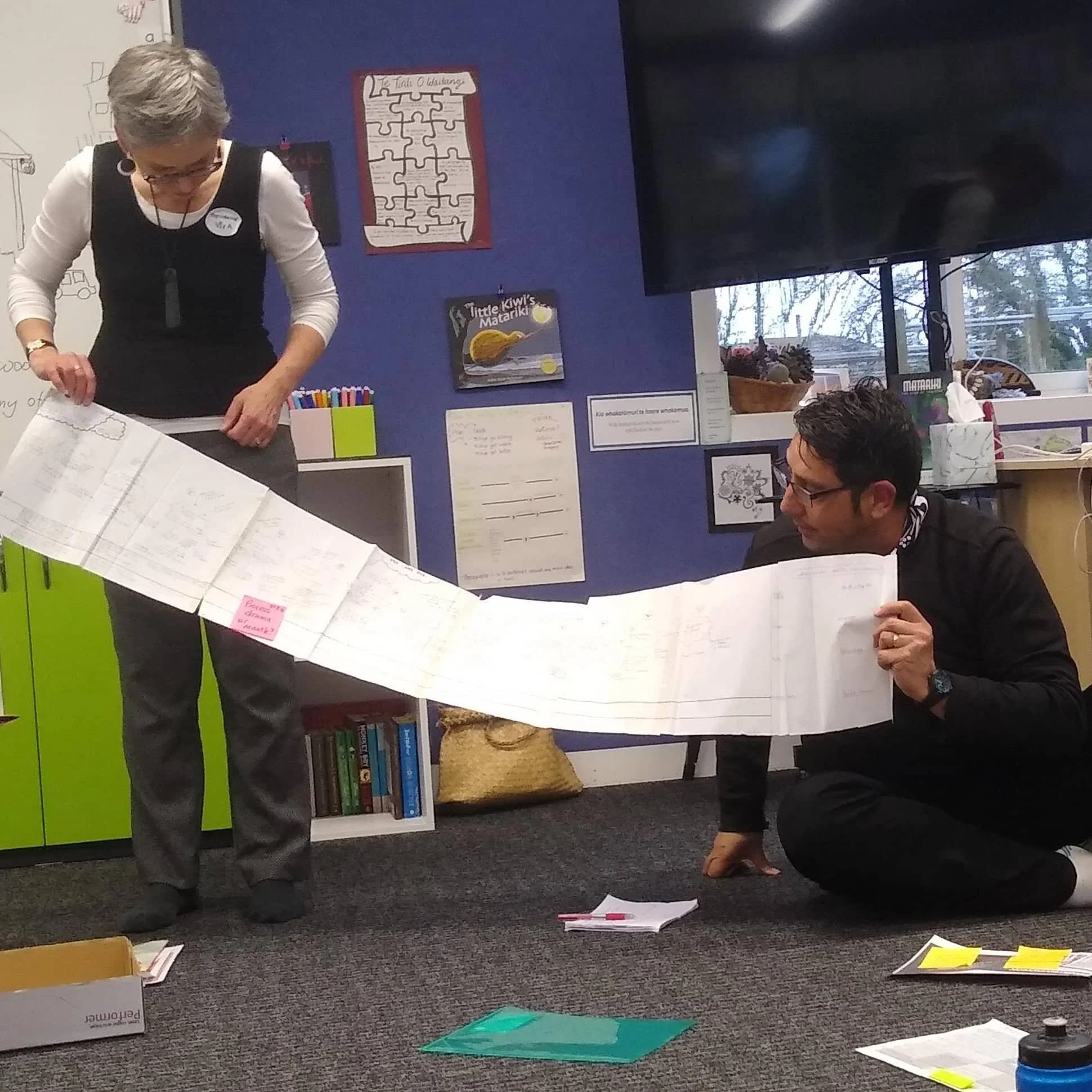 A woman with short gray hair and glasses is standing and looking down, working on a project. She's wearing a white long-sleeve shirt with a black overall and a name tag. There are papers and colored markers on the table in front of her. To her right, a man with glasses and dark hair is sitting on the floor, also working on papers. The background has a blue wall with posters and a large television screen. There are books and items on shelves, and a window with a view outside.