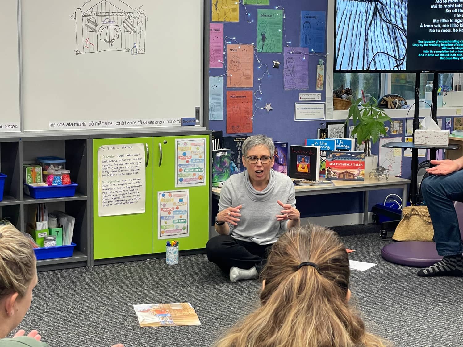 A woman with short gray hair and glasses sitting on the floor in a classroom, speaking to children who are sitting on the carpet. Behind her is a whiteboard, colorful posters, books, and a large TV screen displaying lyrics. The classroom has educational materials and decorations.