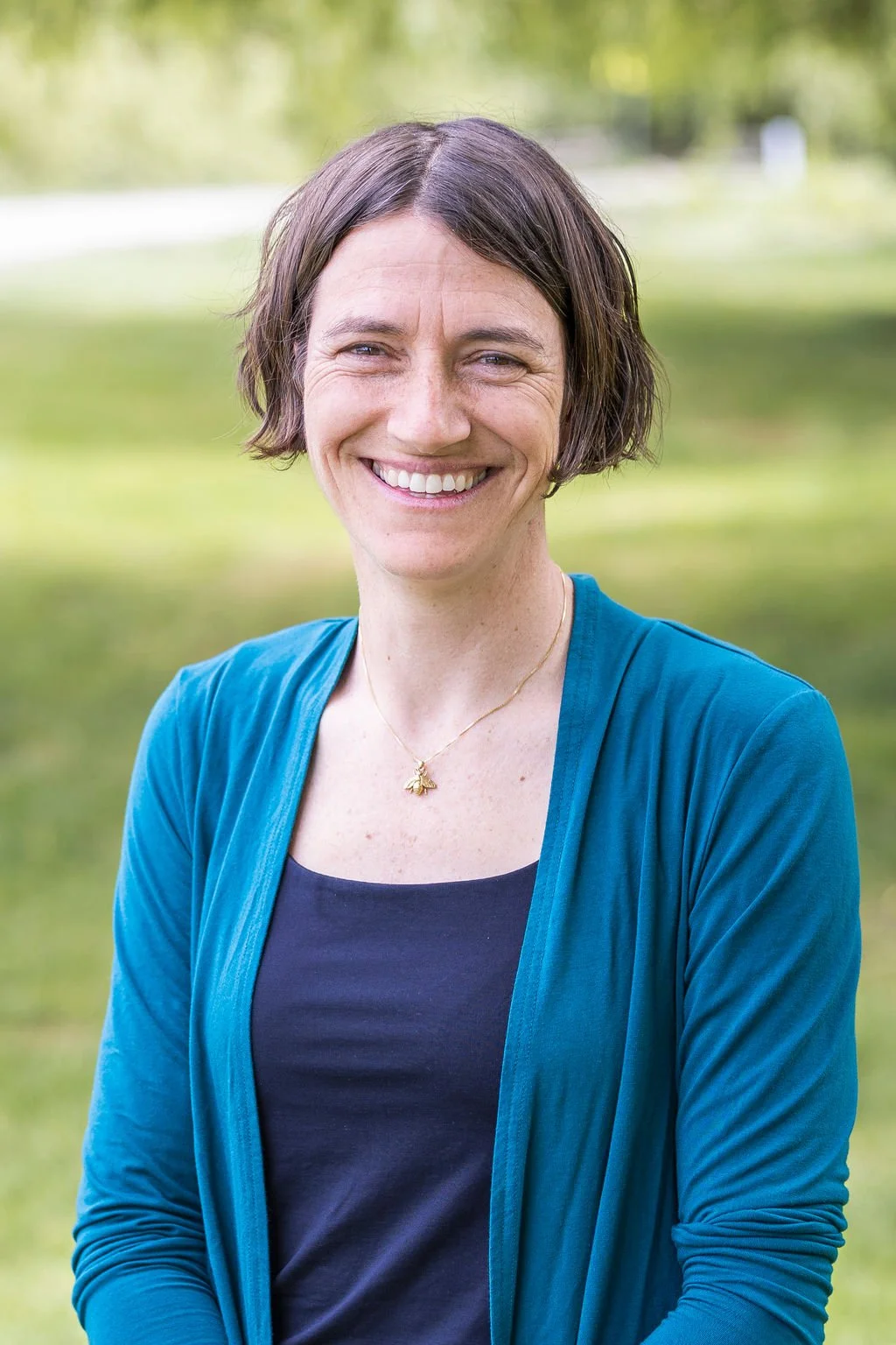 Image of Amy Braunstein smiling in a Blue shirt and blue cardigan against a blurred green background of a park.