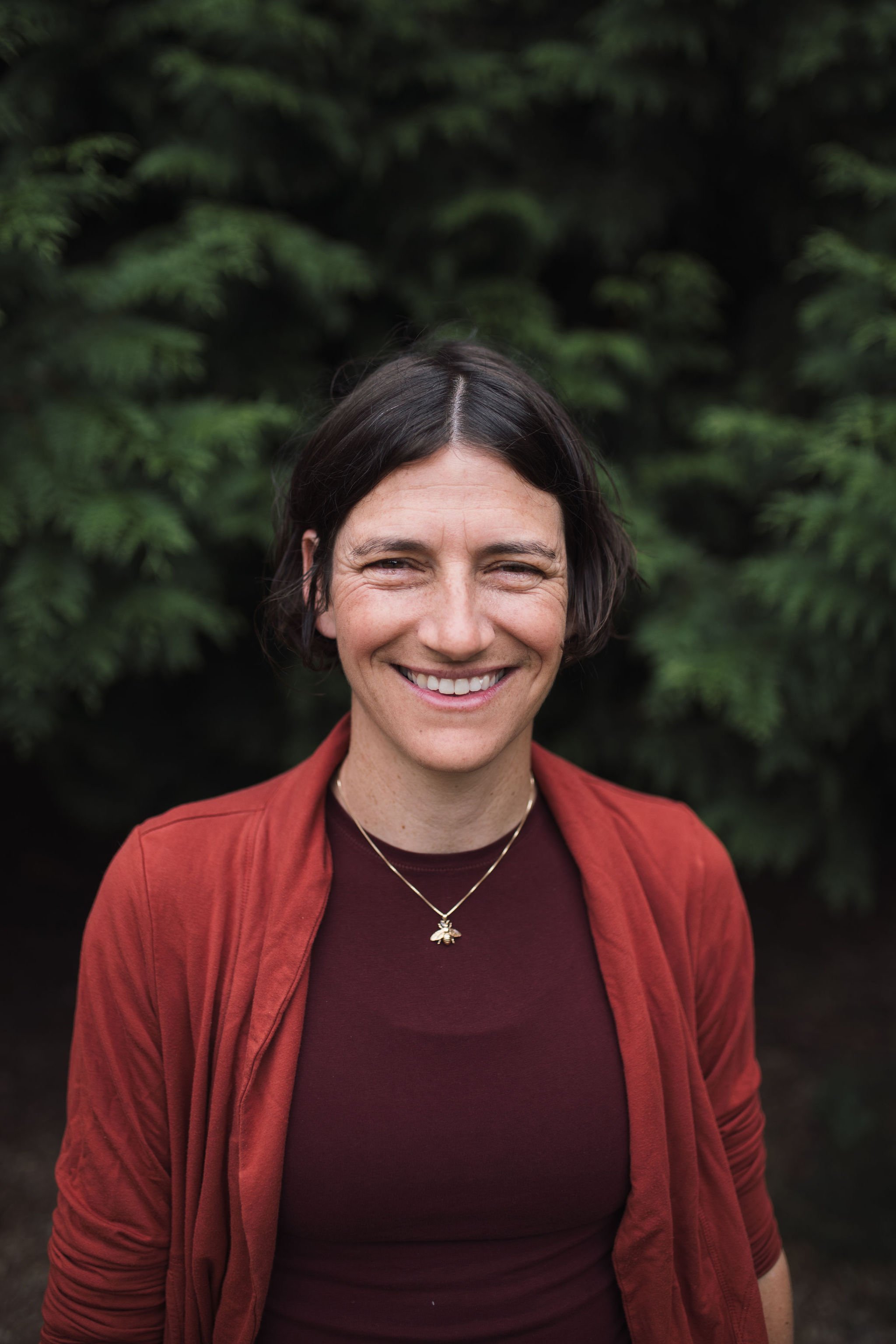 Image of Amy Braunstein smiling in a Blue shirt and blue cardigan against a blurred green background of a park.