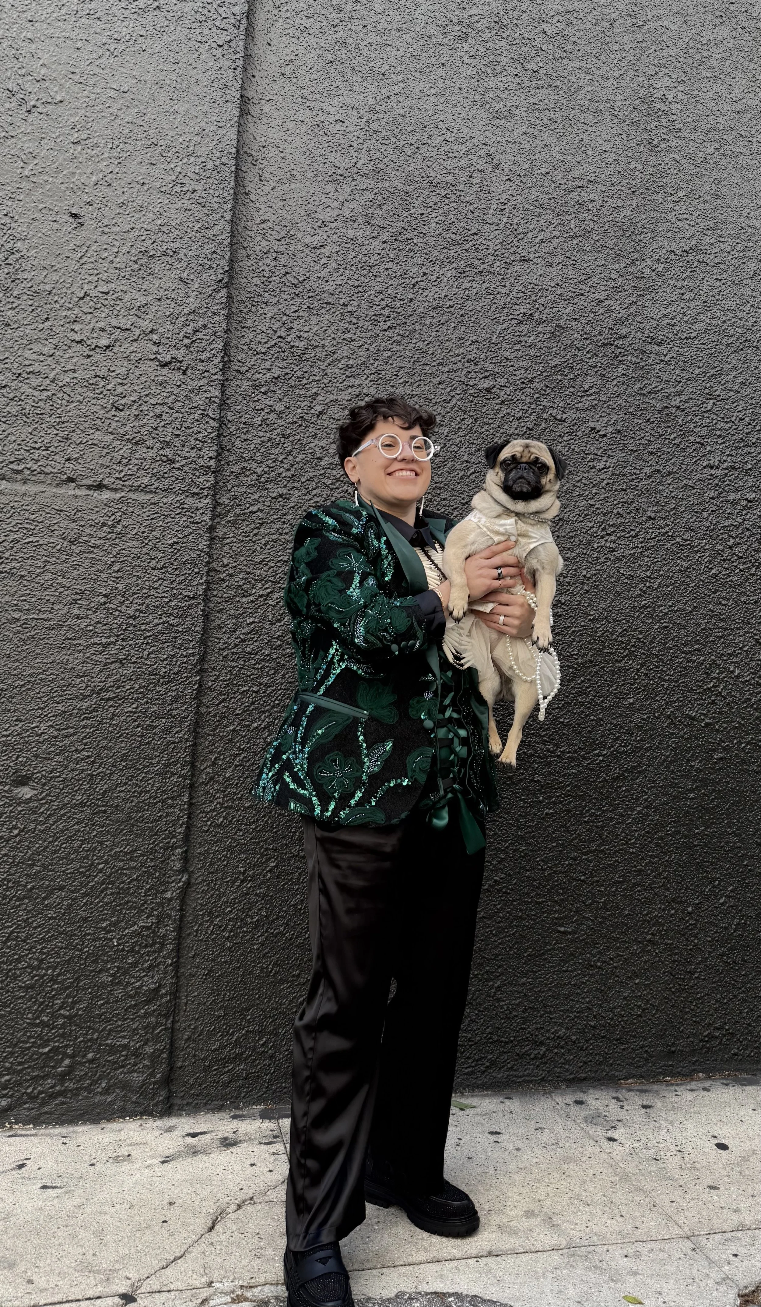 A person in a black suit is giving a kiss to a dog dressed in a white frilly dress at an outdoor wedding, with a bouquet of pink and white flowers to the side.