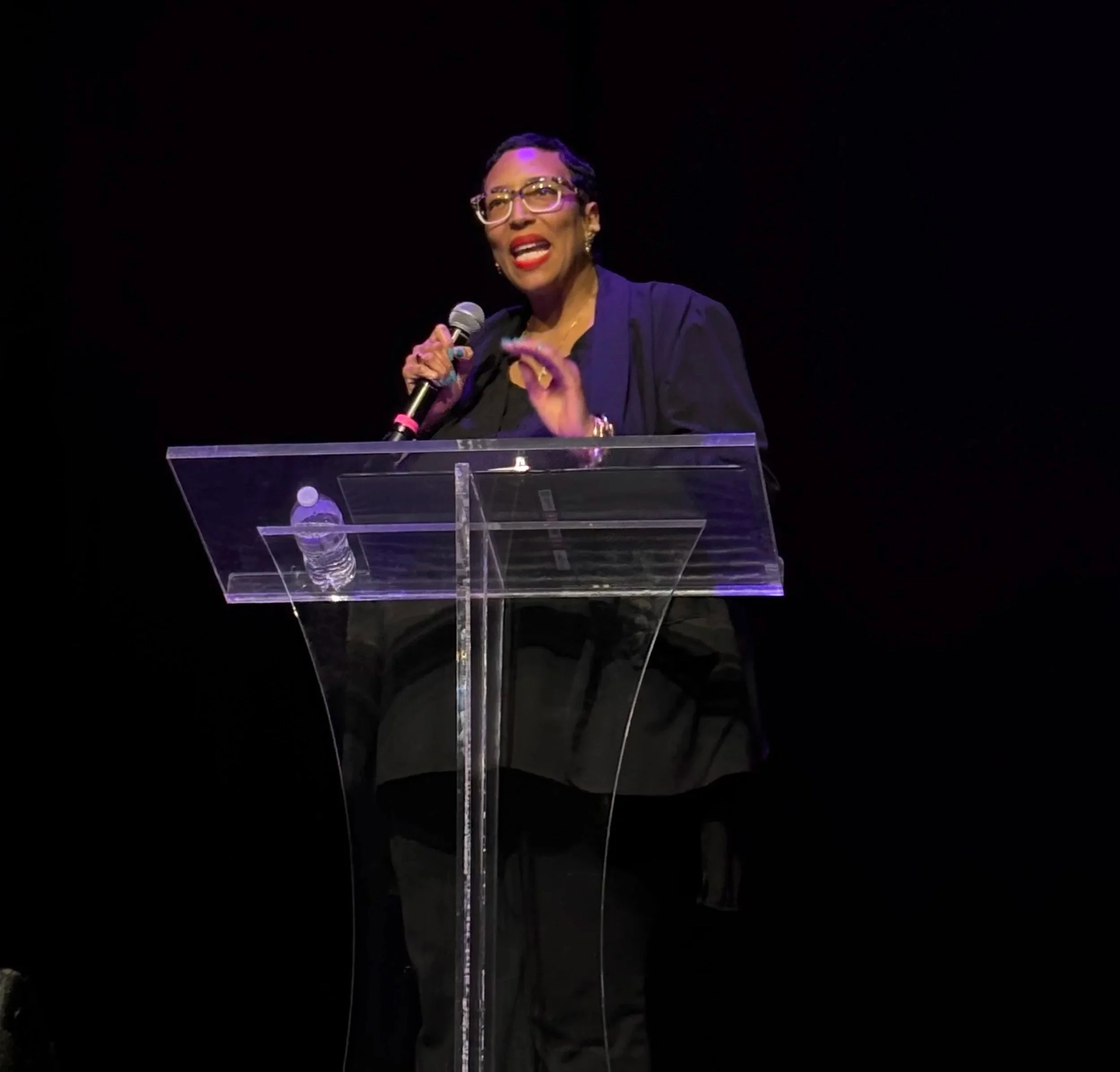 A woman with short hair, glasses, and red lipstick speaking at a podium with a microphone against a dark background.