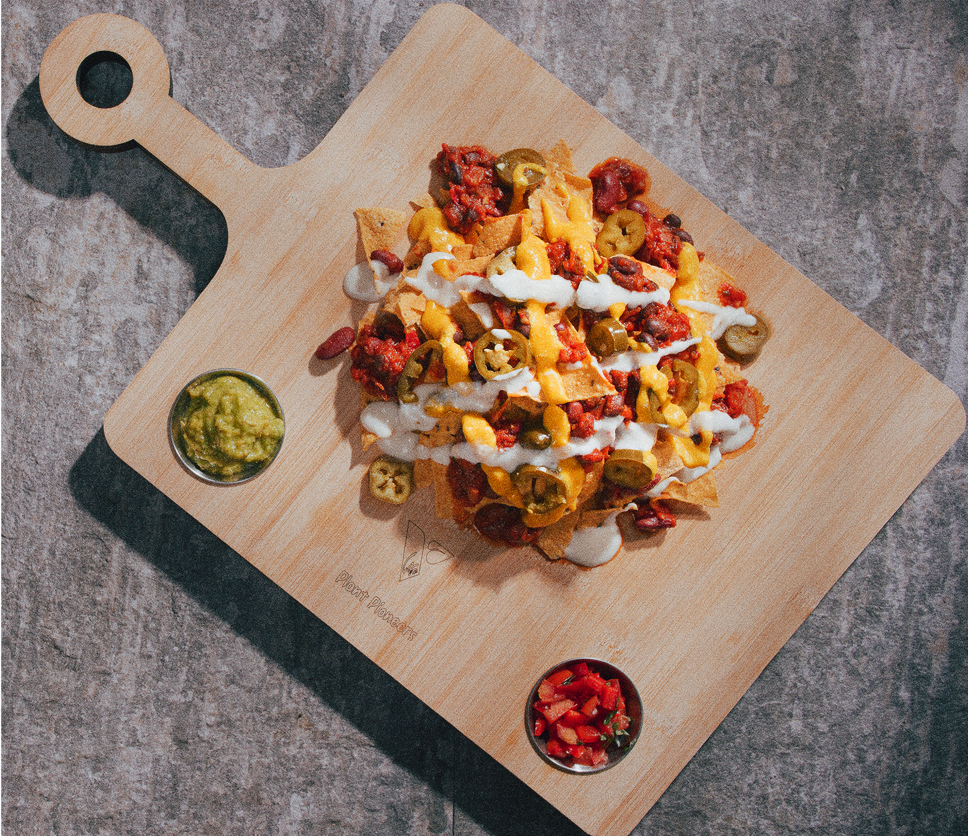 Plate of loaded nachos topped with cheese, jalapenos, sour cream, and salsa on a wooden serving board, with guacamole and salsa in small bowls nearby.