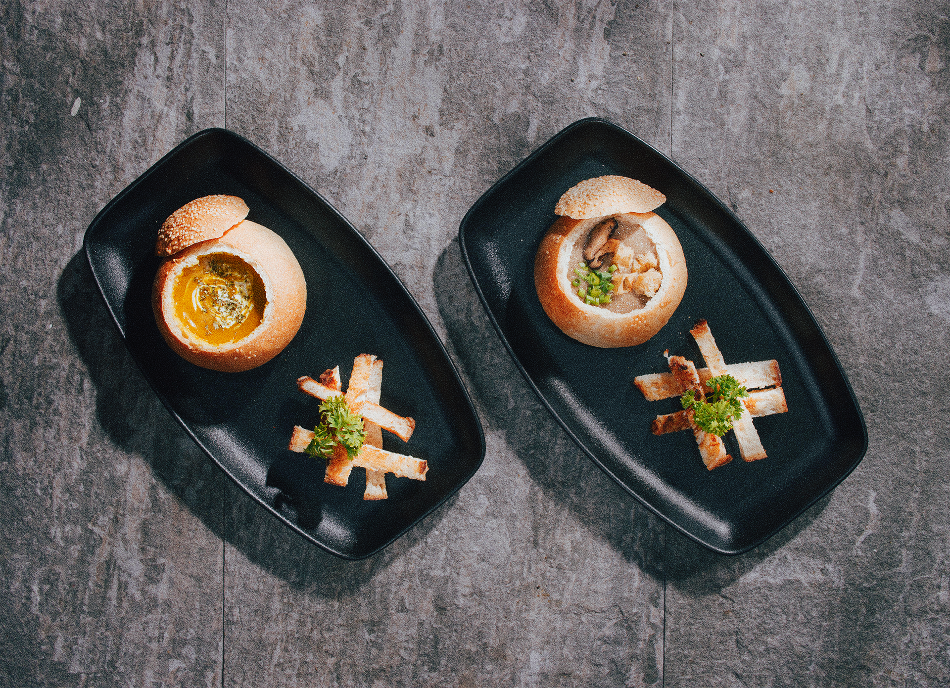 Two black rectangular plates with bread bowls filled with eggs and soup, garnished with green herbs and accompanied by crispy strips of bread on a dark stone surface.