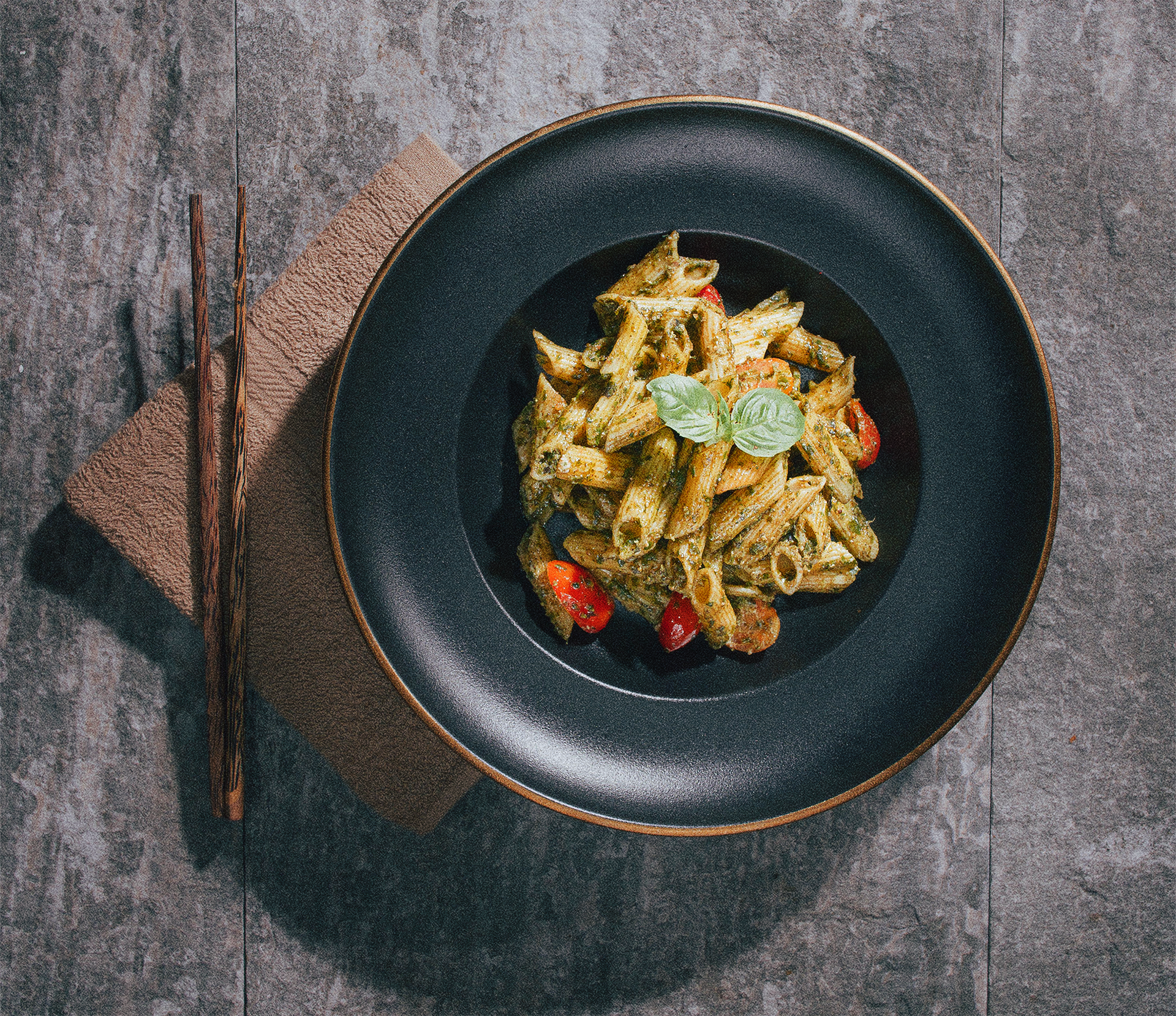 Plate of penne pasta with pesto sauce, cherry tomatoes, and basil leaves on a black plate, with chopsticks resting on a napkin beside it on a gray textured surface.