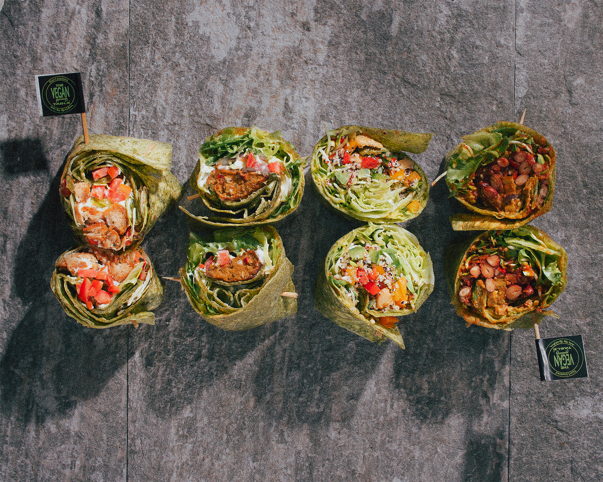 Variety of vegan wraps filled with vegetables, beans, and plant-based protein on a dark slate surface with small vegan flags.