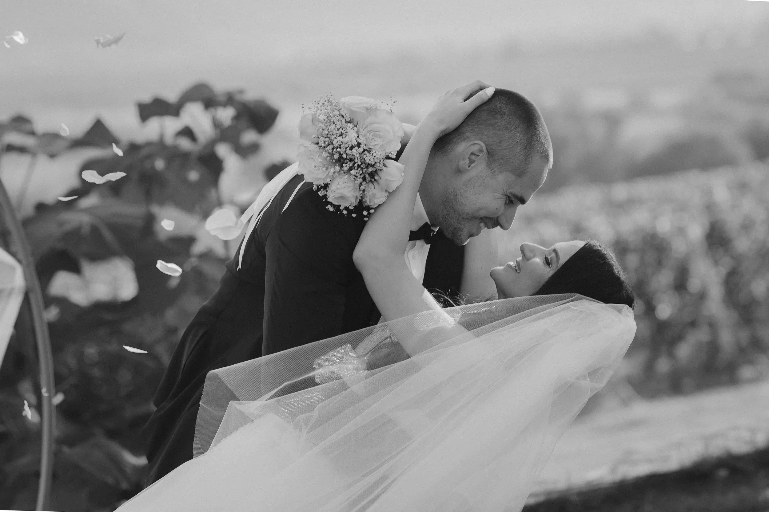 A wedding couple, the groom holding the bride, smiling at each other outdoors with falling flower petals M&ZPhotostory | Bulgaria